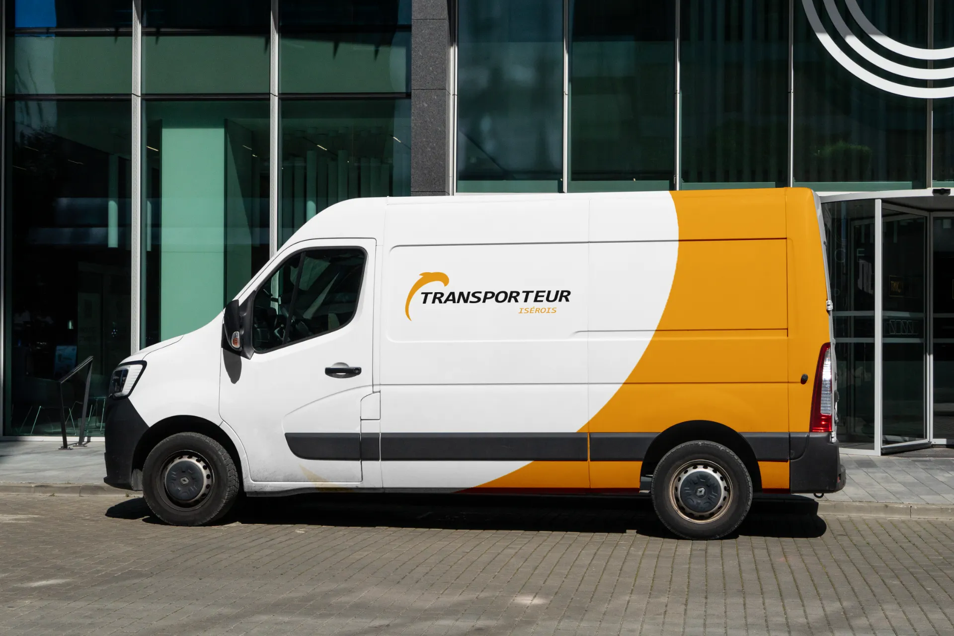 Un fourgon blanc et orange est garé devant un bâtiment moderne avec des fenêtres vertes et un logo "TRANSPORTEUR EXPRESS" sur le côté.