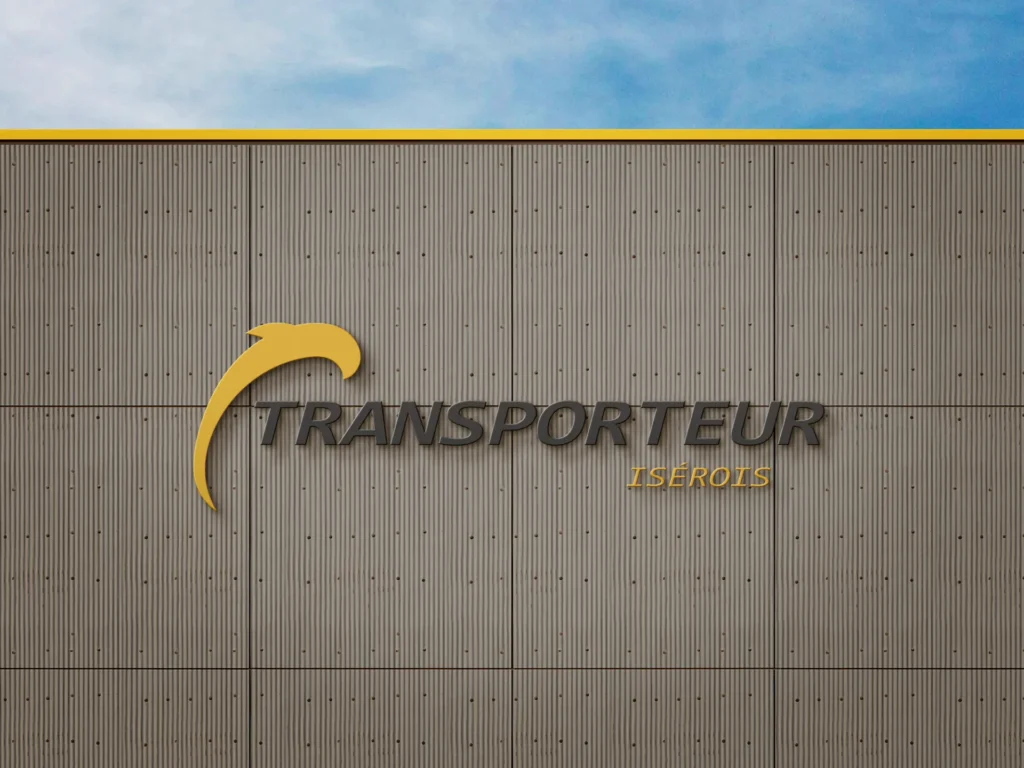 Logo de TRANSPORTEUR ISEROIS sur un mur de bâtiment gris sous un ciel bleu.