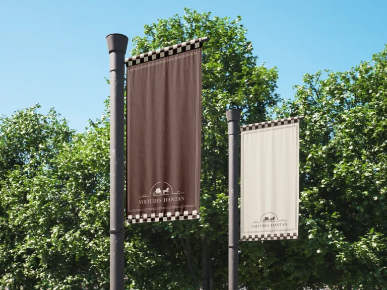 Deux bannières verticales, une marron et une beige, suspendues à des poteaux devant des arbres verdoyants. Elles portent le logo "VOITURES D'ANTAN" et un motif de damiers noirs et blancs.